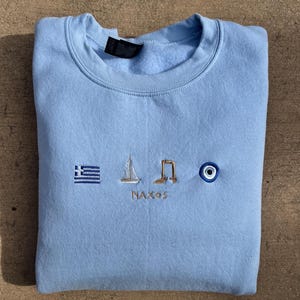 May include: Light blue sweatshirt with embroidered Greek symbols, including a sailboat, a gate, a Greek flag, and an evil eye. The text "Naxos" is embroidered below the symbols.