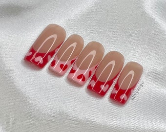 May include: Set of six nude-colored press-on nails with a white outline and a pink and red heart design.