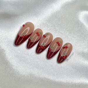 May include: Set of six almond-shaped false nails with a red and nude color gradient. The nails are decorated with small red and green rhinestones.