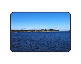 May include: A scenic view of a calm, deep blue ocean under a clear, bright blue sky. Several sailboats are visible on the water, with a treeline and houses in the distance. The image is framed by a black border.