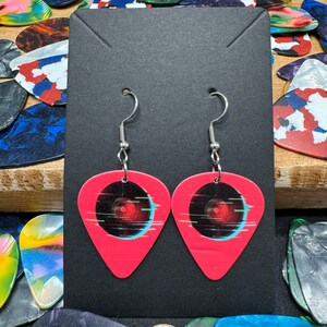 May include: A pair of pink guitar pick earrings with a black and blue record design. The earrings have silver hooks.