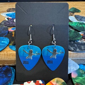May include: A pair of blue guitar pick earrings featuring a picture of a baby swimming underwater. The earrings are silver and have a hook fastening. The guitar picks have the text "Nirvana" printed on them.