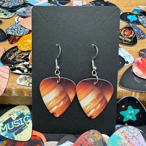 May include: A pair of earrings featuring a guitar pick design with a printed image of a planet. The earrings are silver and have a hook closure.