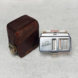 May include: A silver Kodalux light meter with a white diffuser and a leather case. The light meter has a dial with numbers from 2.8 to 30 and a scale with numbers from 5 to 500. The text "DIFFUSOR!" is printed on the diffuser.