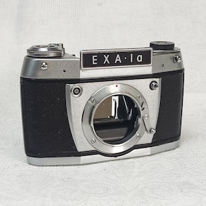 Könnte beinhalten: Eine silberne und schwarze Exa 1a 35mm Filmkamera mit schwarzer Lederimitation. Die Kamera hat ein silbernes Objektiv mit einem roten Punkt oben.