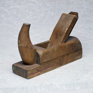 Könnte beinhalten: Ein alter Holzhandhobel mit gebogenem Griff und Klinge. Der Hobel ist aus gealtertem Holz gefertigt und weist deutliche Gebrauchsspuren auf, was auf ein antikes Werkzeug hindeutet. Das Design umfasst eine Klinge und einen gebogenen Griff.