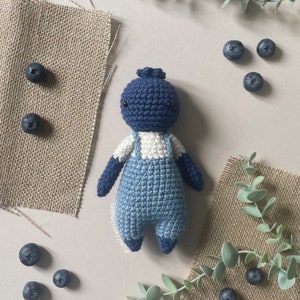 Op de afbeelding: Een gehaakte pop in blauw en wit met een overall. De pop heeft een blauw hoofd met zwarte ogen en een wit gezicht. De pop zit op een lichtbruin oppervlak.