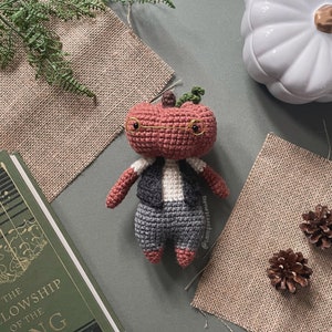 May include: A crocheted pumpkin wearing glasses and a vest. The pumpkin is red and has a green stem. The pumpkin is sitting on a gray surface.