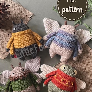 May include: Four crocheted plush creatures with wings, each wearing a different coloured jumper. One is dark grey with a yellow jumper, one is light pink with a blue striped jumper, one is light brown with a green jumper, and one is light green with a red jumper. Text reads PDF pattern.