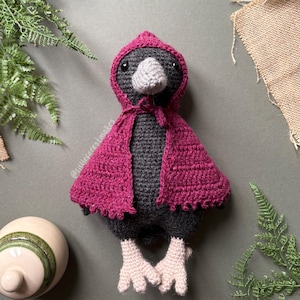 May include: A handmade crochet bird doll with a dark gray body, a gray beak, and light pink feet. The bird wears a magenta hooded cape. The doll is a whimsical, handcrafted item.