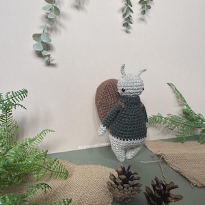 May include: A crocheted snail toy with a brown shell, green sweater, and white body. The snail is standing on a green surface with pine cones and burlap.