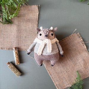 以下が含まれることがあります： 茶色と白の毛皮で編まれた鹿のぬいぐるみで、白いマフラーをしています。ぬいぐるみは茶色の麻布の上に置かれています。