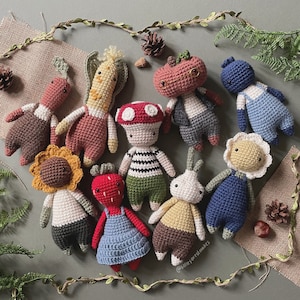 Op de afbeelding: Een verzameling van 10 gehaakte amigurumi-speelgoed, elk met een uniek ontwerp en kleur. De speelgoed omvatten een ​​zonnebloem, een aardbei, een paddenstoel, een pompoen, een wortel, een kabouter, een blauw personage, een wit personage, een geel personage en een bruin personage.