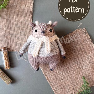 May include: A crochet deer plush toy featuring a brown head with white spots, a light gray sweater, and a cream-colored scarf. The toy has black bead eyes, small antlers, and dark brown hooves. A circular graphic in the corner reads PDF pattern.