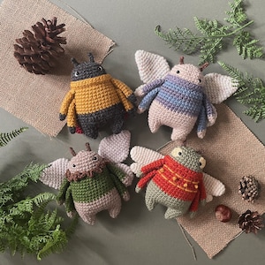 May include: Four crocheted stuffed animal toys in the shape of insects. The toys are in various colours, including green, blue, red, and yellow. The toys are sitting on a brown hessian surface.