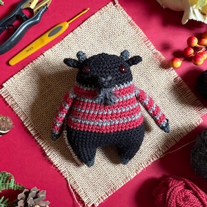 Op de afbeelding: Een handgemaakte zwarte gehaakte geit met een rood-grijze gestreepte trui. De geit heeft kleine hoorns, een grijze baard en rode ogen. Hij zit op een vierkant van jute tegen een rode achtergrond. Knutselspullen staan op de achtergrond.