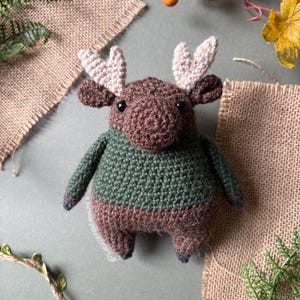 May include: A handmade crochet moose toy with a brown body, green sweater, and cream-colored antlers. The moose has small black eyes and is posed on a neutral background with burlap and greenery accents. The toy is approximately 6 inches tall.
