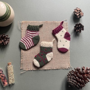 Puede incluir: Tres calcetines de Navidad en miniatura de ganchillo en verde, blanco y burdeos con diferentes patrones. Los calcetines se muestran sobre una superficie de arpillera.