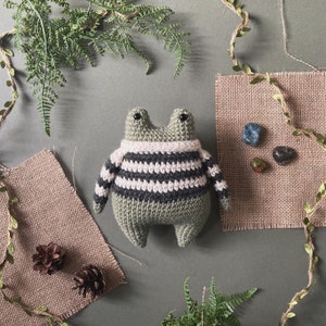 Könnte beinhalten: Ein grüner gehäkelter Frosch, der einen weiß-schwarz gestreiften Pullover trägt. Der Frosch sitzt auf einer braunen Jutestruktur.