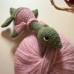 May include: A handmade crochet turtle in shades of green and pink rests on a ball of pink yarn. The turtle has a detailed head, small limbs, and a shell-like covering. The text "@littleforest makes" is visible.