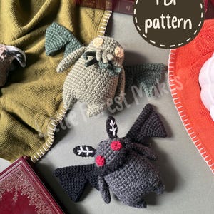 May include: Two crocheted plush toys: one light green creature with wings and tentacles, and one dark gray creature with red eyes, white ear markings, and wings. A circular brown graphic with the white text PDF pattern is visible.