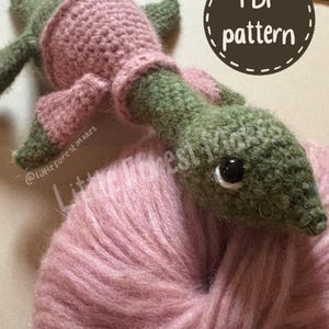 May include: A green crocheted creature with a long neck and pink clothing resting on a ball of pink yarn. The image includes a circular graphic with the text PDF pattern and the watermark text @little forest makes visible on the image.