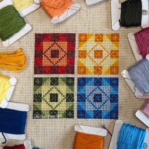 Könnte beinhalten: Ein Kreuzstich-Sampler mit vier Quadraten, jedes mit einem geometrischen Muster in Rot, Gelb, Grün und Blau. Der Sampler ist von Spulen farbigen Stickgarns umgeben. Der Text "Little Forest Makes 2026" ist sichtbar.