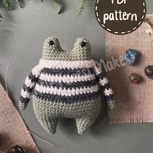 Peut inclure: Une peluche grenouille crochetée avec un corps vert sauge et un pull à rayures horizontales blanches et gris anthracite foncé. La grenouille a deux petits yeux en perles noires et une tête ronde. Une étiquette brune affiche PDF pattern.
