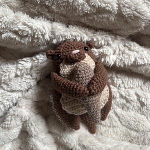 May include: A handmade crochet otter in shades of brown and beige. The otter is hugging itself, with a pink nose and black eyes. The plush toy is on a soft, white, textured surface. The text "littleforestmakes" is visible.
