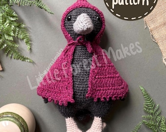 Op de afbeelding: Een gehaakte kraaienpop van donkergrijs garen, met een lichtgrijze snavel en lichtroze voeten. De kraai draagt een donker magenta cape met capuchon die bij de nek is gestrikt. Een bruine badge met witte tekst PDF pattern. De tekst Little Forest Makes is zichtbaar.