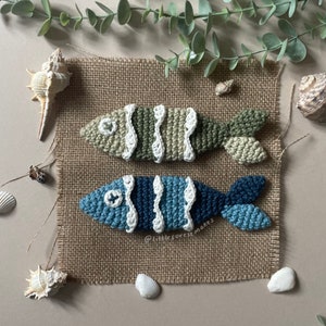 Pode incluir: Dois enfeites de peixe de crochê em um fundo de juta. Os peixes são feitos com fio verde, azul e branco. Os peixes têm um padrão ondulado e olhos brancos.