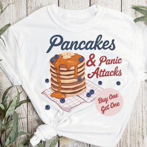 Könnte beinhalten: Weißes T-Shirt mit einem Grafikdruck von einem Stapel Pfannkuchen mit Blaubeeren und Sirup sowie dem Text "Pancakes & Panic Attacks" und "Buy One Get One".