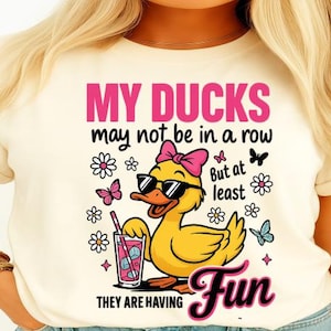 Peut inclure: T-shirt crème avec un dessin d'un canard jaune portant des lunettes de soleil et un nœud rose. Le texte dit "MY DUCKS may not be in a row But at least THEY ARE HAVING Fun" avec des fleurs et des papillons.