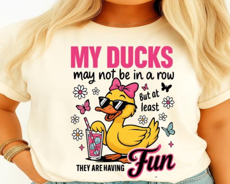May include: Cream-colored t-shirt with a graphic of a yellow duck wearing sunglasses and a pink bow. The text reads "MY DUCKS may not be in a row But at least THEY ARE HAVING Fun" with flower and butterfly illustrations.