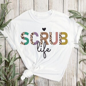 May include: White t-shirt with the text "SCRUB life" in colorful letters with a leopard print pattern. The letters are outlined in black.