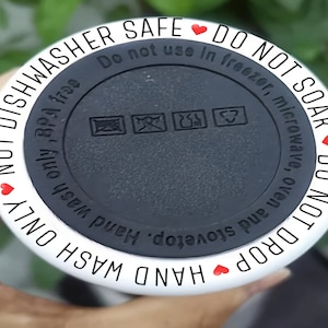 May include: White ceramic mug with black text that reads "NOT DROP HAND WASH ONLY NOT DISHWASHER SAFE DO NOT SOAK DO NOT USE IN FREEZER, MICROWAVE, OVEN AND STOVETOP. HAND WASH ONLY BPA FREE". The mug has a black lid with a recycling symbol and other symbols.