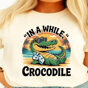 May include: Cream-colored t-shirt featuring a cartoon crocodile wearing sunglasses and a Hawaiian shirt. The design includes the text "IN A WHILE, CROCODILE" in black and white, with a sunset and palm trees in the background.