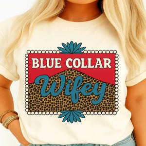May include: Cream-colored t-shirt featuring the text "BLUE COLLAR Wifey" in a retro design. The text is set against a red background with a leopard print pattern and teal accents. The shirt has short sleeves.