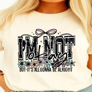 以下が含まれることがあります： クリーム色のTシャツ。黒字で「I'M NOT lazy BUT IT'S ALL GONNA BE ALRIGHT」と書かれ、パステルカラーの花柄のデザイン。ピンクのリボンと星も含まれています。