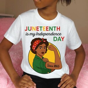 May include: White t-shirt with the text "Juneteenth is my Independence Day" in red, green, and black. The shirt features a graphic of a woman flexing her arm, with the words "Since 1865" on her arm.