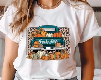 Può includere: Maglietta bianca con una grafica di un camioncino color verde acqua pieno di zucche e girasoli. Il camioncino ha la scritta "Pumpkin Farm" sul lato. La grafica è su uno sfondo leopardato.