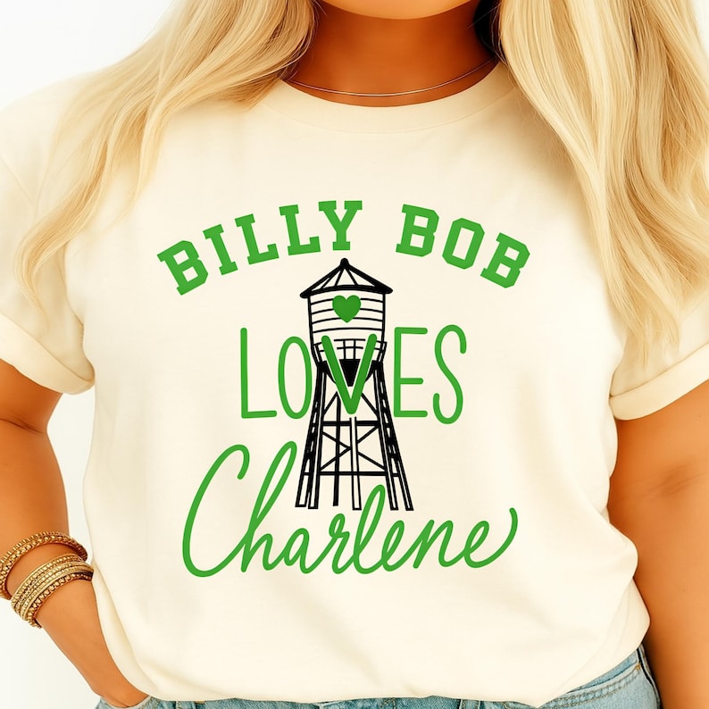 May include: Cream-colored t-shirt with the text "BILLY BOB LOVES Charlene" in green, featuring a black and white water tower graphic with a green heart. The shirt has short sleeves.