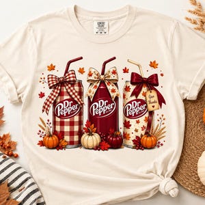 Op de afbeelding: Een crèmekleurig T-shirt met korte mouwen en een afbeelding van drie Dr. Pepper-blikjes versierd met strikken, rietjes en herfstelementen. De blikjes zijn omringd door kleine pompoenen, rode bessen en herfstbladeren. Een label bevat de tekst Give Thanks.