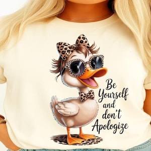 Puede incluir: Camiseta color crema con un pato de dibujos animados que lleva gafas de sol, un lazo y una pajarita, todos con un diseño de estampado de leopardo. El texto "Be Yourself and don't Apologize" está escrito en guion negro.
