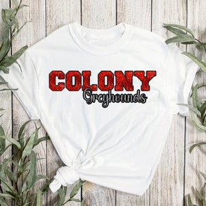 May include: White t-shirt with the words "COLONY Greyhounds" in red glitter lettering. The word "COLONY" is in a bold, block letter style, and "Greyhounds" is in a script font. The shirt is displayed on a light wood background with green foliage.
