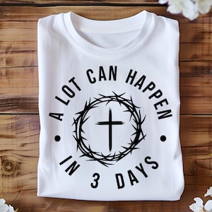 May include: White t-shirt with a black graphic. The graphic features a cross inside a circle of thorns, with the text "A LOT CAN HAPPEN IN 3 DAYS" in a circular arrangement around the cross.