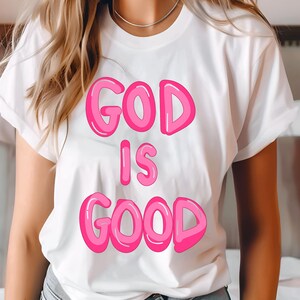 May include: White t-shirt with the text "GOD IS GOOD" in bubble-style pink letters with a darker pink outline. The shirt has short sleeves and is being worn by a person.