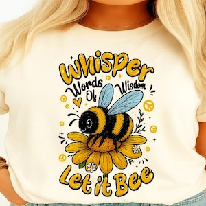 Puede incluir: Camiseta color crema con una abeja de dibujos animados sobre una flor amarilla. La abeja tiene rayas negras y amarillas y alas azules. El texto "Whisper Words of Wisdom Let it Bee" está en amarillo y negro.
