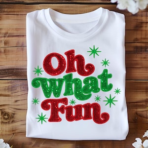 May include: White t-shirt with the phrase "Oh What Fun" in red and green glitter lettering. Green starbursts surround the text. The shirt is folded on a wooden surface.