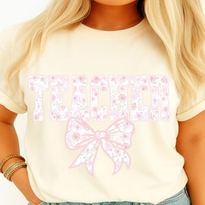 Może przedstawiać: Kremowy t-shirt z napisem "TEACHER" w dużych, jasnoróżowych kwiatowych literach. Pod tekstem znajduje się pasująca różowa kwiatowa kokarda. Koszulka ma krótkie rękawy i okrągły dekolt.